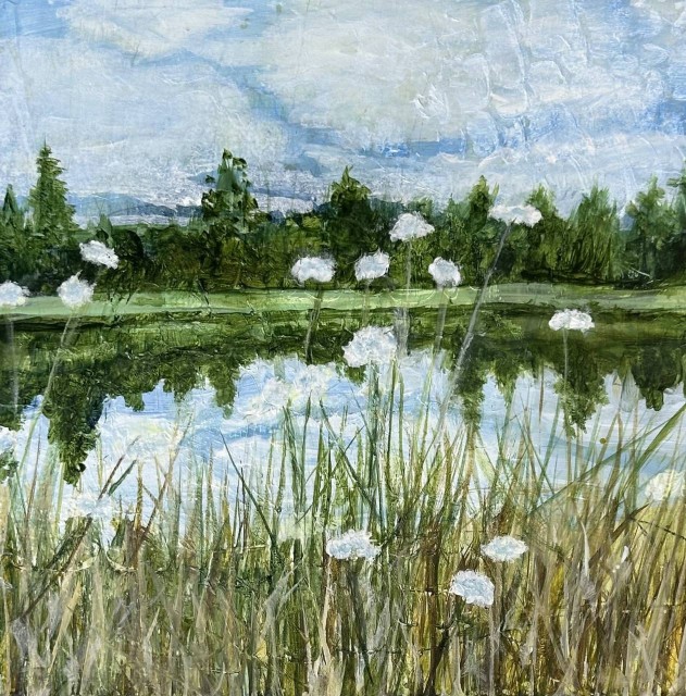 This is an acrylic painting of a riverbank landscape along a river's smooth, flat water. Tall grasses are in the foreground, with Queen Anne's Lace, tall-stemmed flowers mingled in with the grasses. Across the river is a row of trees beyond a flat grass area. The trees are reflected clearly in the river; the water mirrors the sky and trees. By Juanita Bellavance